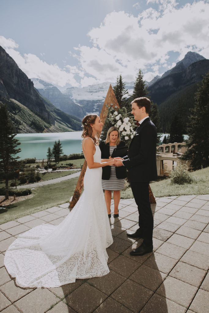 Couple stands holding hands in front of wedding arch and officiant overlooking Lake Louise at wedding venue Fairmont Chateau Lake Louise