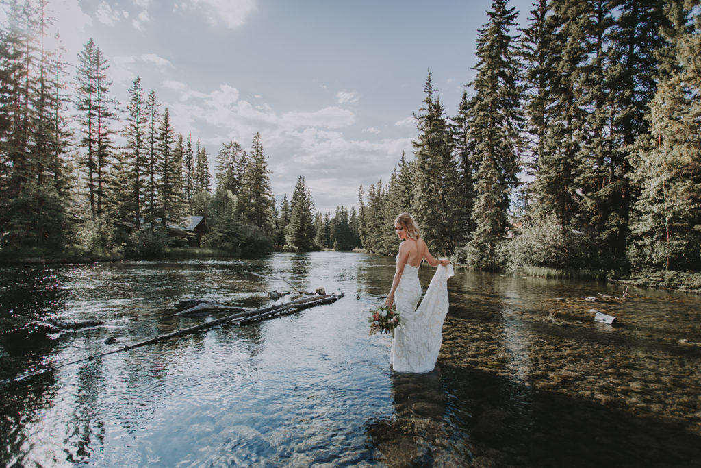 Bride holds the bottom of her wedding gown in one hand and a bouquet in the other as she stands in the middle of a river surrounded by evergreen trees