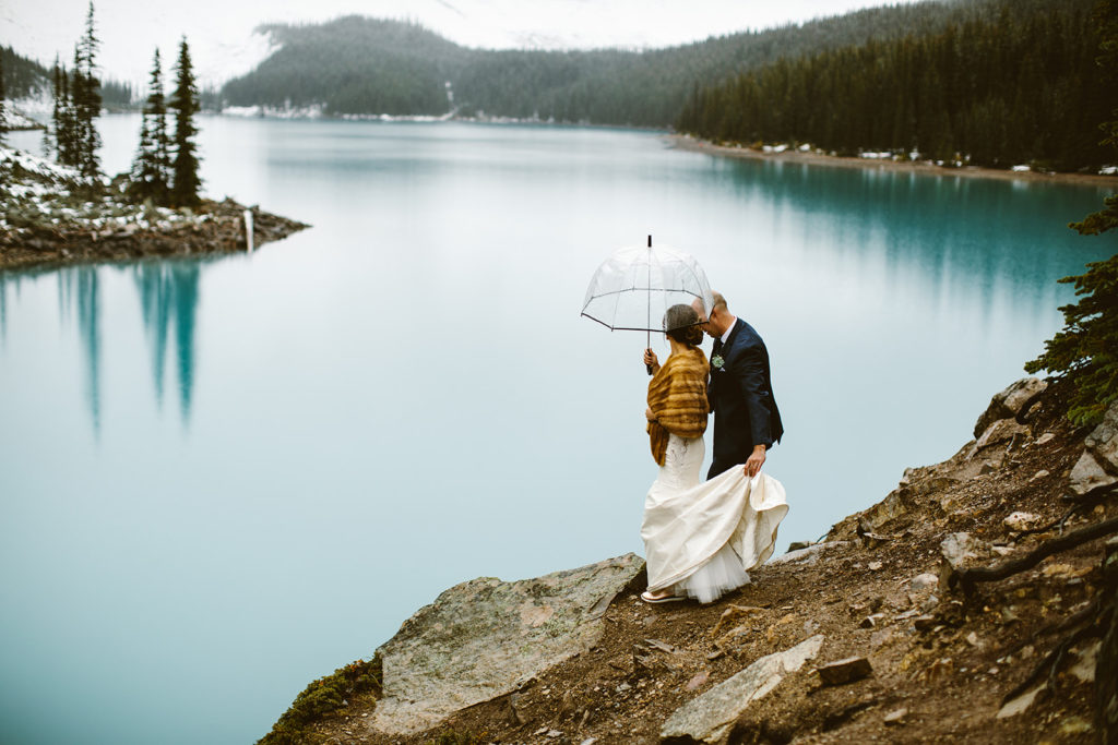 Couple stands on a cliff overlooking a blue lake, huddled together under a clear umbrella.