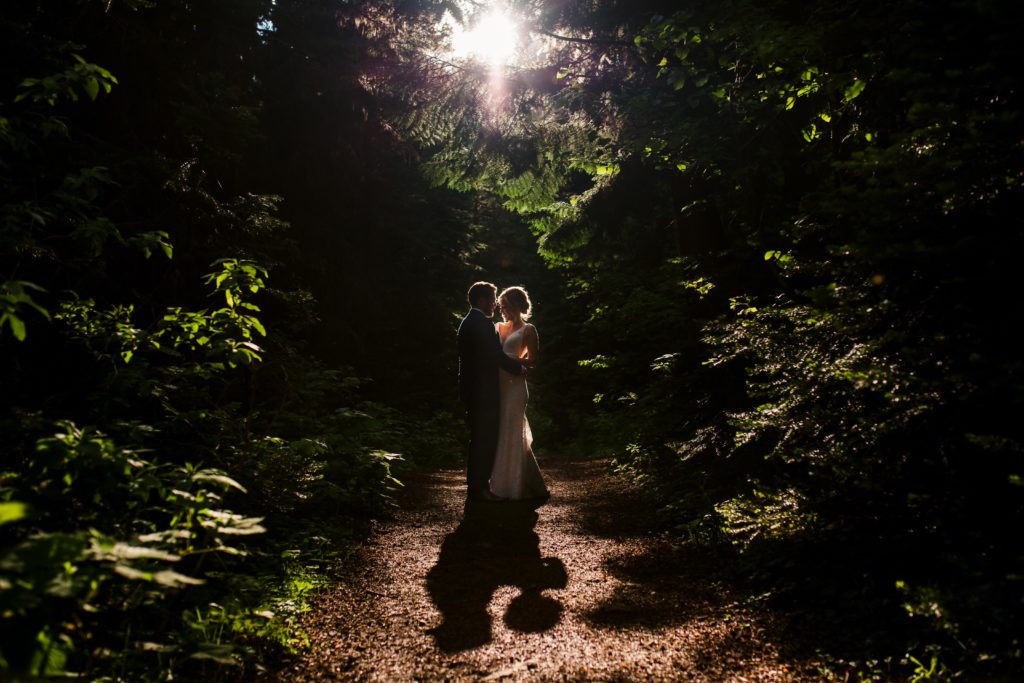 A bride and groom surrounded by lush forest embrace in their summer wedding while sunlight peeks through above