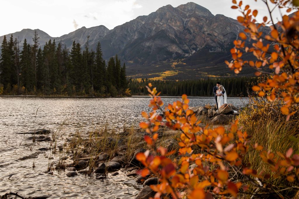 Autumn leaves in foreground with wedding couple embracing in the background standing at the edge of a lake, yellow larch trees seen in the treeline of the mountains